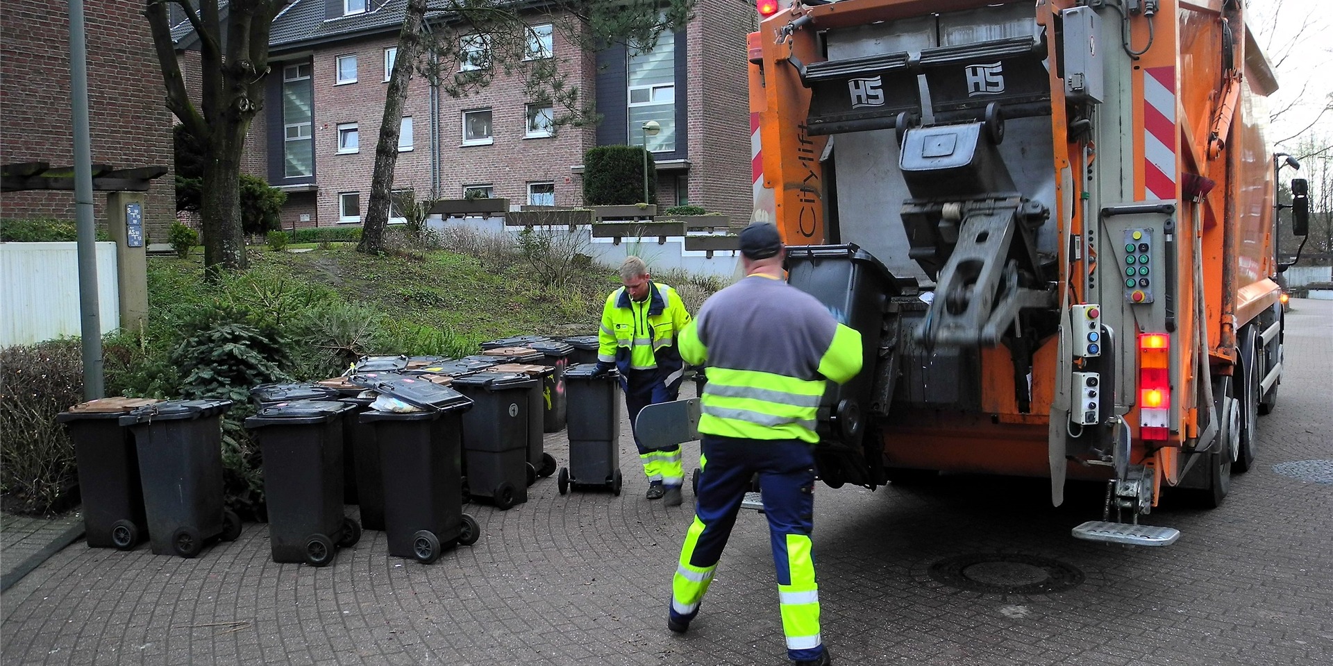 Zwei Mitarbeiter der Müllabfuhr stehen hinter einem Müllwagen.