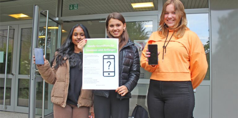 Die Schülerinnen Tharsha Thirukumaran, Elina Neumann und Leonie Neumeyer stehen vor dem Eingang des Waltroper Gymnasiums und halten Plakate und Handys in die Kamera.