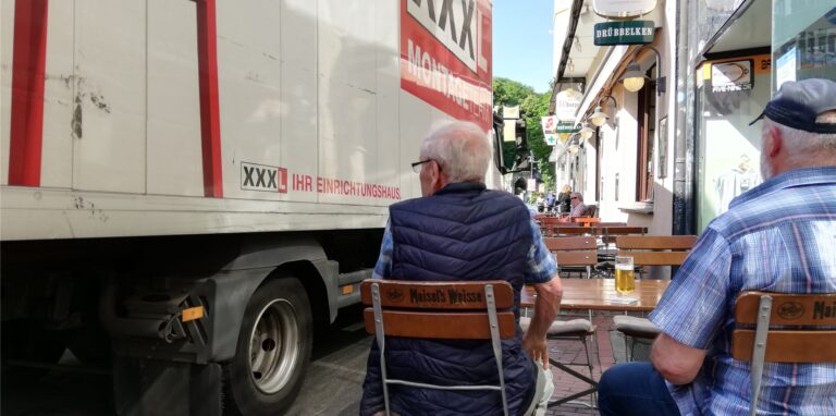 Ein Lastwagen fährt ganz nah an einem Senior vorbei, der neben einem weiteren alten Mann bei einem Bier draußen an einem Tisch vor einer Gaststätte sitzt.
