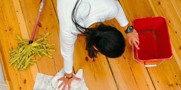 Eine Frau wischt mit der Hand einen Holzfußboden, neben ihr steht ein roter Eimer.