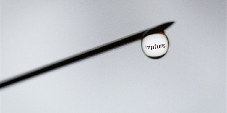 Das Wort „Impfung“ ist in einem Tropfen an der Nadel einer Spritze zu sehen.