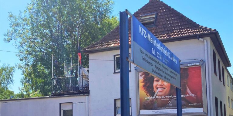 Ein weißes Haus mit einem rußgeschwärzten Balkon, im Vordergrund befindet sich eine Werbetafel.