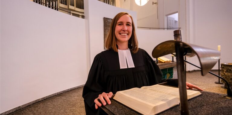 Eine junge Pfarrerin steht in einer Kirche am Redepult, auf dem eine Bibel liegt.