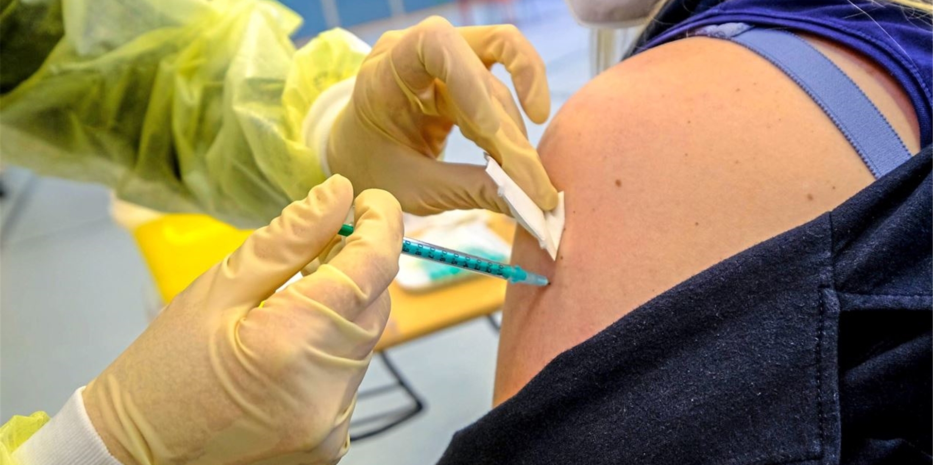 Nahaufnahme einer Frauenschulter, in die jemand mit Schutzhandschuhen eine Spritze mit Impfstoff setzt.