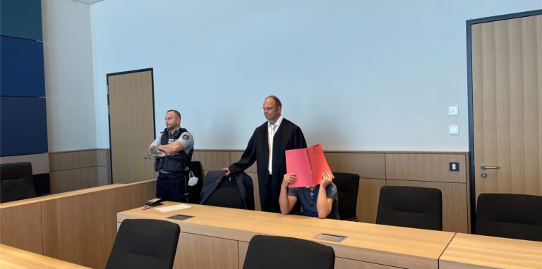 Drei Männer in einem Gerichtssaal, einer in der Uniform eines Justizbeamten, einer in Verteidiger-Robe, ein dritter sitzend mit einem Schnellhefter vor dem Geischt.