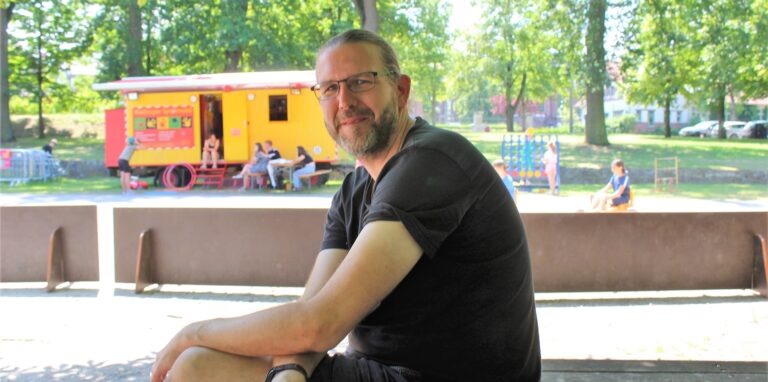 Ein Mann sitzt vor dem Hintergrund des Kinderferienspaßes Waltrop auf einer Bank.