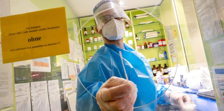 Ein Mitarbeiter einer Apotheke hält ein Teststäbchen für einen Corona-Schnelltest durch die Öffnung einer Scheibe.