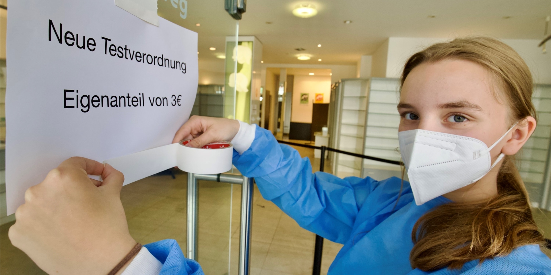 Die Mitarbeiterin eines Testzentrums klebt an eine Glaswand ein Schild auf dem steht, dass Coronatests künftig einen Eigenanteil von 3 Euro kosten.