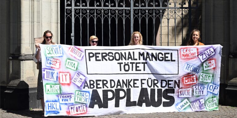 Streikende Pflegekräfte von Unikliniken halten ein Transparent mit der Aufschrift „Personalmangel tötet – Aber danke für den Applaus“.