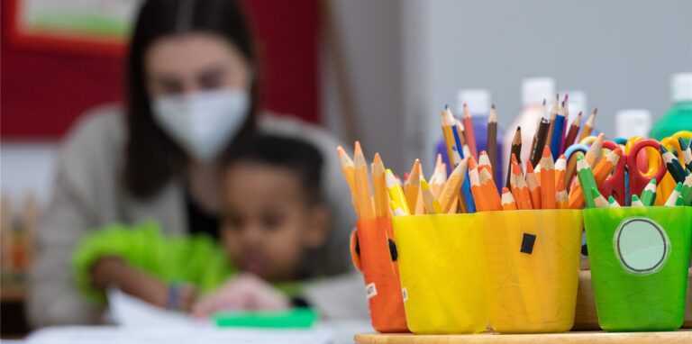 Eine Erzieherin, die eine medizinische Maske trägt, sitzt mit einem Kind in einer Kita hinter Bechern mit Buntstiften. Der hessische Elternbund fordert landesweit einheitliche Quarantäne-Regeln in Kitas.