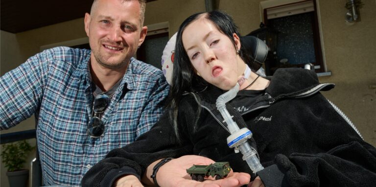Dustin Raatz kniet neben dem Rollstuhl von Chantal vor deren Haus und zeigt ein Modell seines tonnenschweren Bundeswehrfahrzeugs MAN Kat 1.
