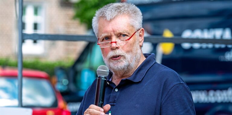Ein Mann mit Brille, Bart und grauen Haaren hält ein Mikrofon in der Hand.