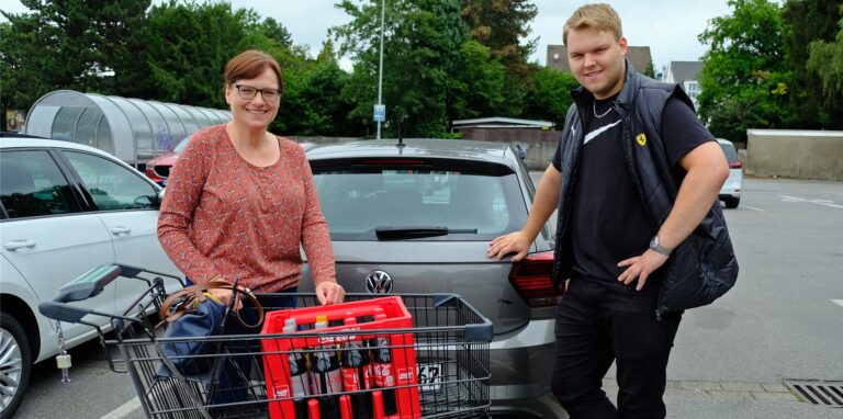 Eine Frau und ein junger Mann stehen mit einem Einkaufswagen auf einem Parkplatz vor einem Auto.