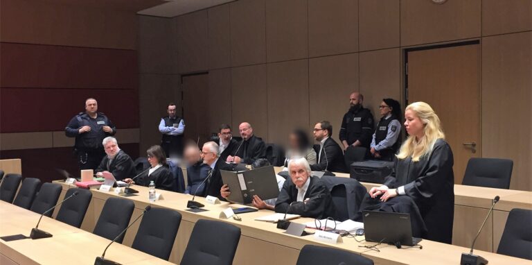 Verteidiger, Justizbeamte und unkenntlich gemachte Angeklagte sitzen in einem Gerichtssaal.