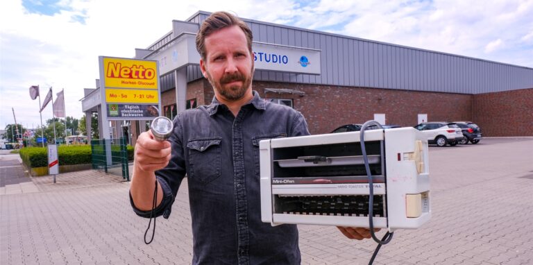 Ein Mann hält vor einer Netto-Filiale einen Toaster und eine Taschenlampe in den Händen.