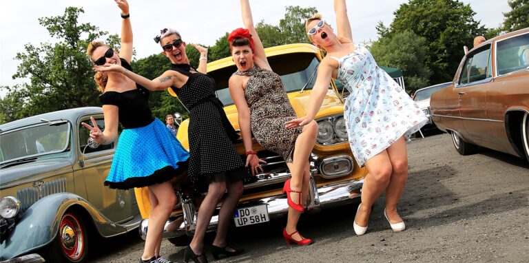 Vier Frauen in Outfits der 50er-Jahre posieren vor einem Auto.