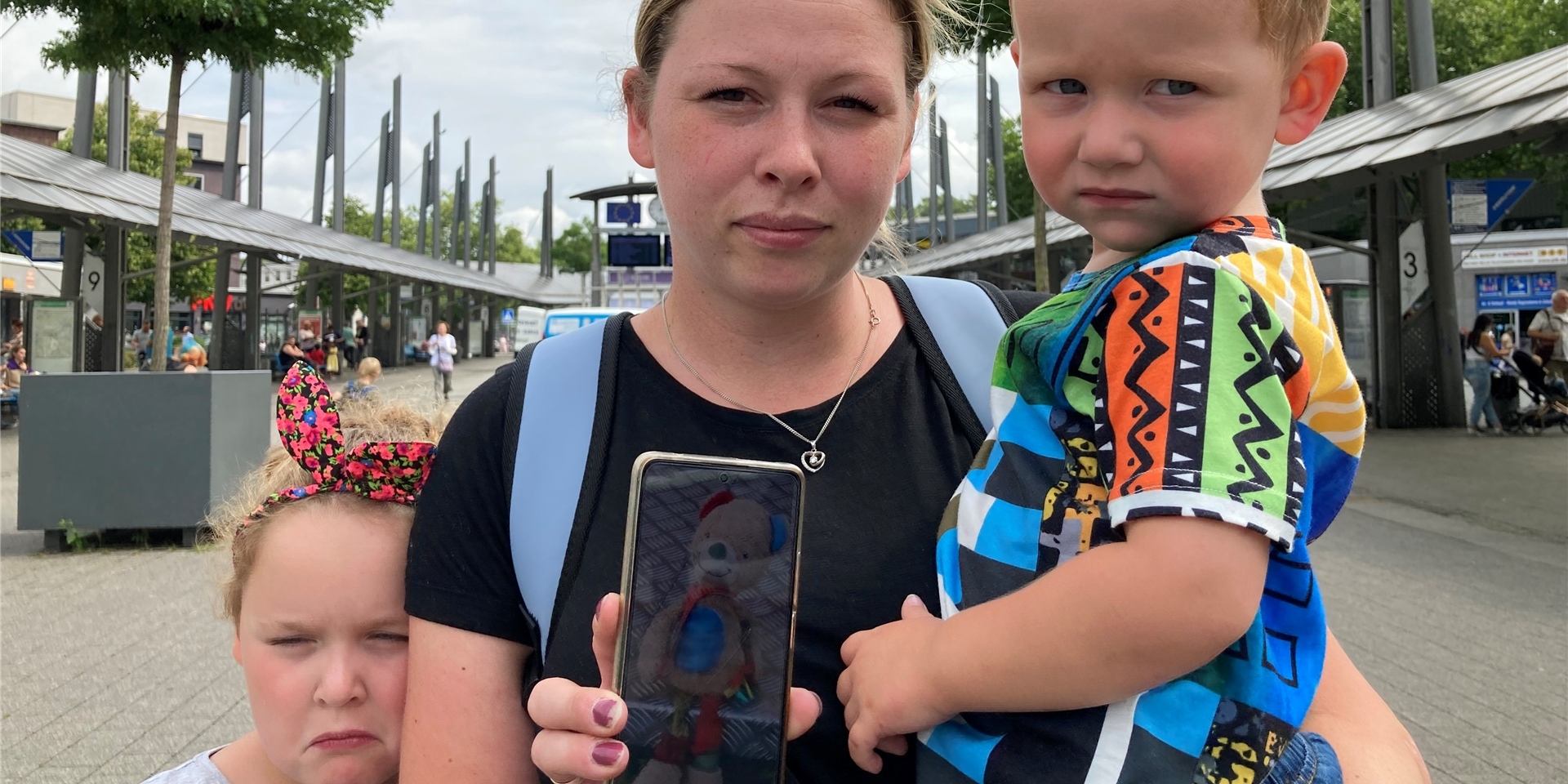 Mila (9), Mutter Nina und Ben Wischnewski aus Recklinghausen stehen am Busbahnhof. Mutter Nina hält ihr Smartphone in die Kamera, auf dem Display ist das Foto eines Teddybären zu sehen.
