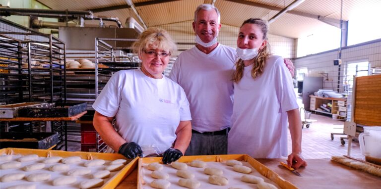 In einer Backstube stehen zwei Frauen und ein Mann vor Backblechen mit Brötchen-Teiglingen.