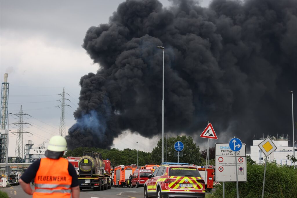 Einsatzfahrzeuge der Feuerwehr stehen unweit einer Zufahrt zum Chemiepark, über dem eine dunkle Rauchwolke aufsteigt. Am 27. Juli 2021 war es in Leverkusen zu einer verheerenden Explosion gekommen.