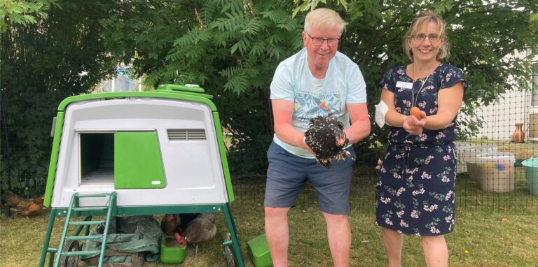 Ein Mann steht mit einem Huhn in der Hand neben einem kleinen, mobilen Stall. Daneben präsentiert eine Frau ein Ei.