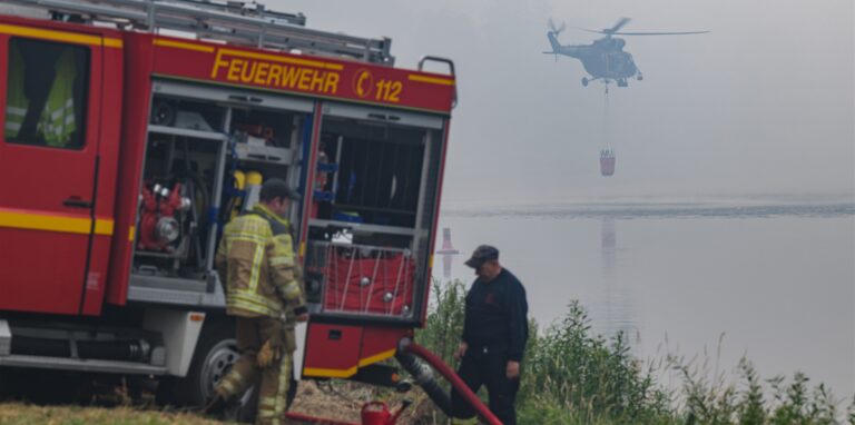Ein tschechischer Militärhubschrauber holt mit einem Löschwasser-Außenlastbehälter Wasser aus der Elbe während deutsche Feuerwehrleute am Elbufer an der Löschwasserversorgung im Kampf gegen die Waldbrände im Nationalpark Sächsische Schweiz arbeiten.