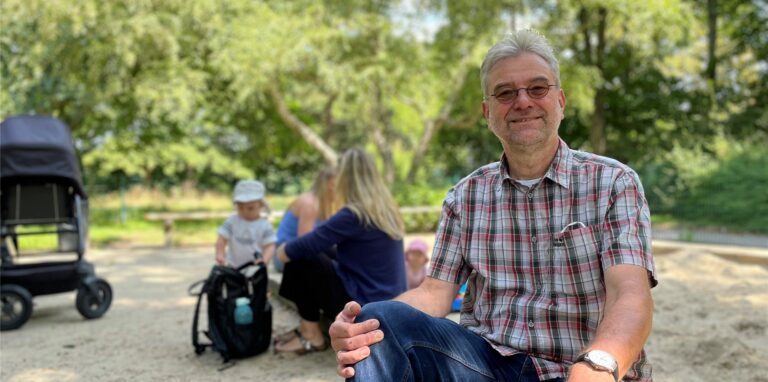 Matthias Fritsch, Mitarbeiter der Stadt, sitzt auf an einem Sandkasten. Im Hintergrund zwei Mütter mit ihren Kleinkindern, die im Sandkasten an der Hallenbadwiese in Castrop-Rauxel spielen.