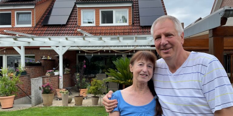 Ingrid Klinger und Jörg Potowski stehen in ihrem Garten. Im Hintergrund sind die Terrasse und das Dach mitsamt der PV-Anlage zu sehen.