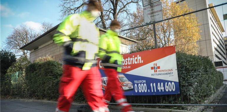 Zwei Rettungssanitäter gehen auf einer Straße an einem Plakat vorbei (Symbolfoto).