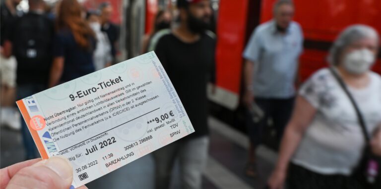 Ein 9-Euro-Ticket für Juli 2022 wird am Hauptbahnhof hochgehalten, während Zugreisende aus einer Regionalbahn aussteigen.