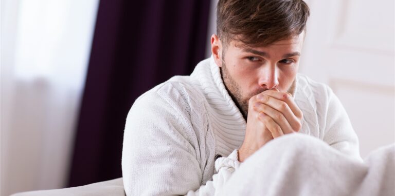 Ein Mann sitzt frierend mit einer Decke auf dem Sofa und bläst in seine Hände