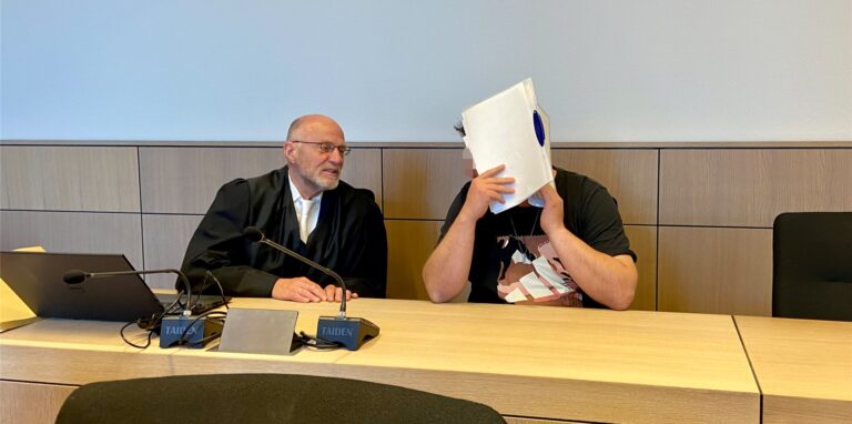 zwei Männer sitzen in einem Gerichtssaal, einer hält sich eine Mappe vors Gesicht