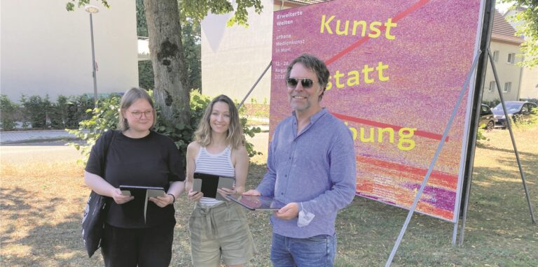 Startpunkt der Ausstellung „Erweiterte Welten“ ist die große Werbetafel an der Dr.-Erich-Klausener-Straße in Marl-Hüls. (v.r.) Marls Museumsdirektor Georg Elben, Medienkünstlerin Anastasija Delidova und Luise Klonowski, Volontärin am Skulpturenmuseum, testen, ob die Technik funktioniert.