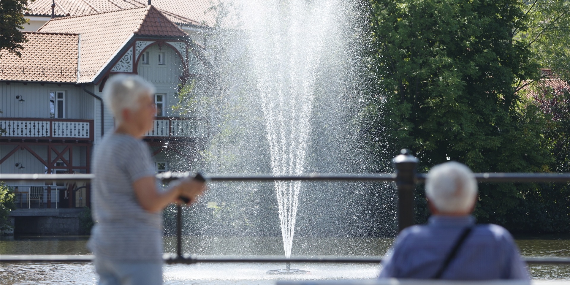 Besucher sitzen auf einer Bank vor einer Wasserfontäne im Forellenteich.