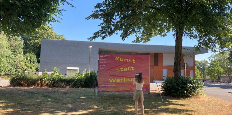 Anastasija Delidova steht vor der Plakatwand gegenüber dem Skulpturenmuseum in Marl-Hüls, wo der Ausstellungsparcours „Erweiterte Welten“ beginnt.