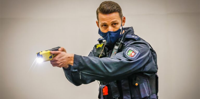 Ein Polizist hält einen Elektro-Taser in der Hand und hat eine Bodycam an seiner Schulter befestigt.