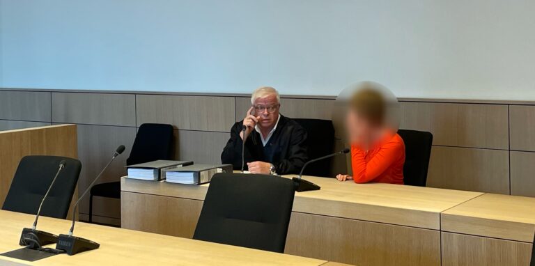 Ein Verteidiger in schwarzer Robe und eine unkenntlich gemachte Angeklagte in rotem Oberteil sitzen in einem Gerichtssaal.
