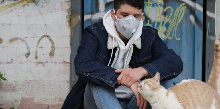 Ein junger Mann sitzt mit einer Corona-Schutzmaske, streichelt eine Katze und schaut traurig vor sich hin.