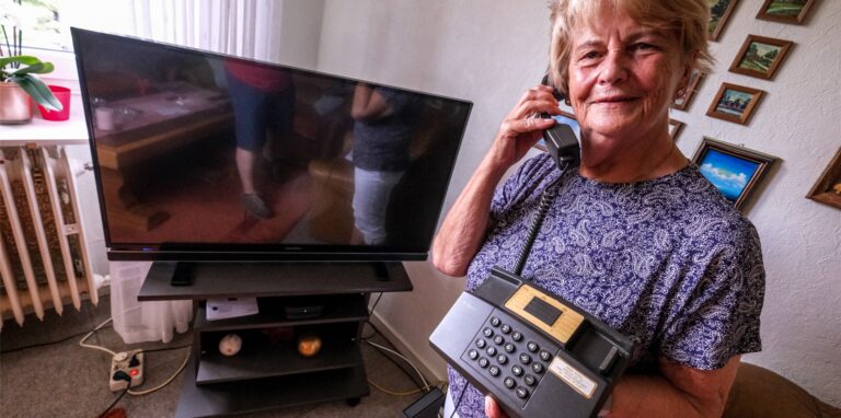 Erika Kubig aus dem Nordviertel in Recklinghausen steht in ihrem Wohnzimmer. Hinter ihr steht ein ausgeschalteter Fernseher, in der linken Hand hält sie ein Telefon, in der rechten Hand einen Telefonhörer.