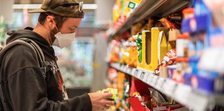 Ein Kunde steht in einem Supermarkt vor einem Regal, und trägt dabei einen Mundschutz.