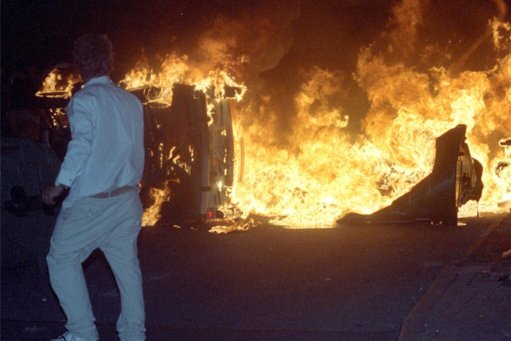 Ein Mann steht am 27.08.1992 vor einem brennenden Pkw auf einer Straße am zentralen Asylbewerberheim von Mecklenburg-Vorpommern in Rostock-Lichtenhagen. Ein Mann steht am 27.08.1992 vor einem brennenden Pkw auf einer Straße am zentralen Asylbewerberheim von Mecklenburg-Vorpommern in Rostock-Lichtenhagen.