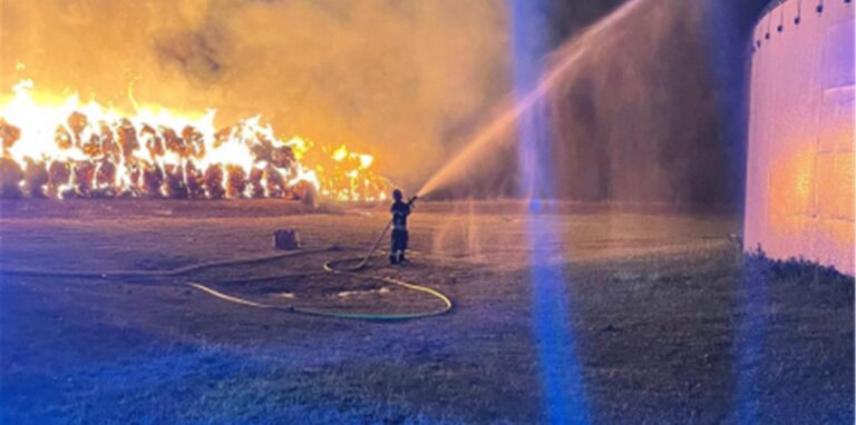 Ein Feuerwehrmann steht mit einem Löschschlauch auf einem Feld. Im Hintergrund sind brennende Strohballen.