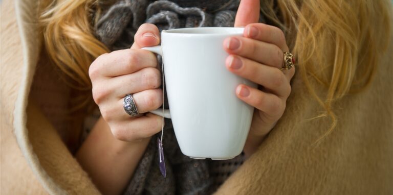 Eine Frau hat eine Decke um sich und wärmt die Hände an einer Tasse Tee.