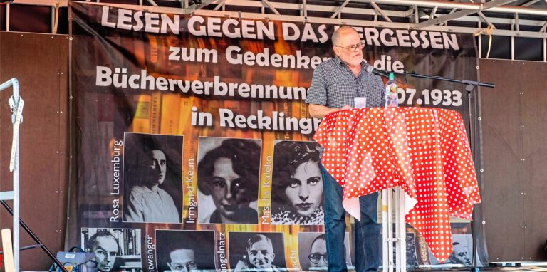 Ein Mann steht auf einer Bühne an einen Bistrotisch und spricht in ein Mikrofon. Im Hintergrund hängt ein Plakat mit der Aufschrift „Lesen gegen das Vergessen“.