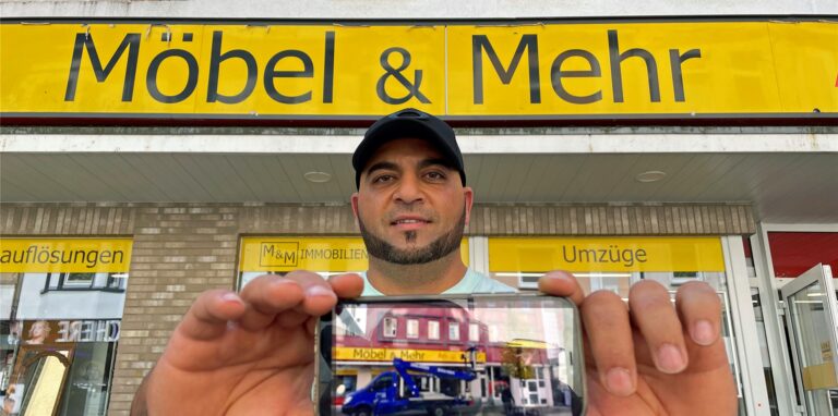 Immobilienbesitzer Yesser Falfoul zeigt sein Smartphone in die Kamera. Auf dem Display ist ein Foto des unrenovierten Hauses hinter ihm zu sehen.