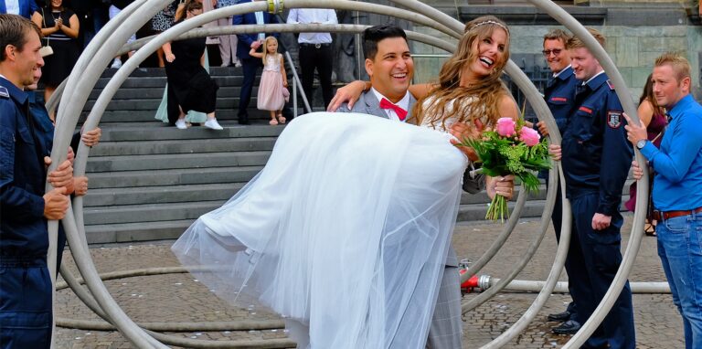 Ein Hochzeitspaar vor dem Rathaus in Recklinghausen. Er trägt sie auf Händen. Männer in Feuerwehr-Uniform stehen mit Schläuchen Spalier.