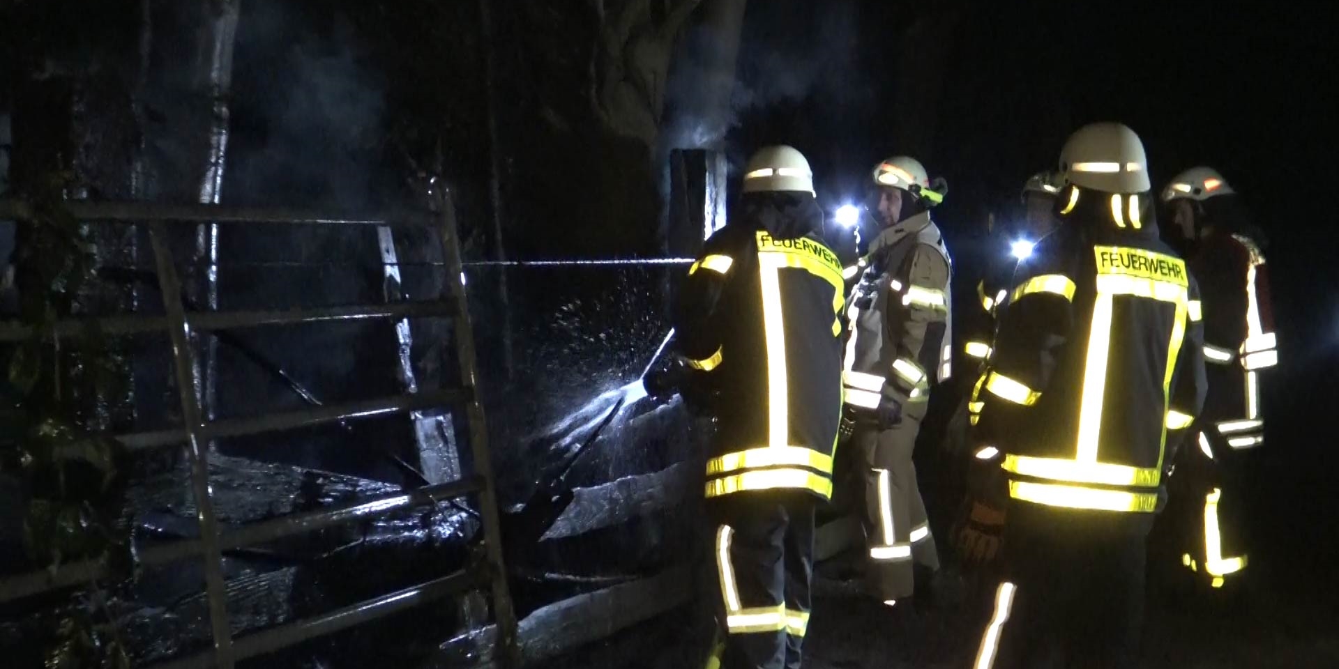Mehrere Feuerwehrleute stehen vor einem Zaun und spritzen mit einem Schlauch Wasser auf qualmende Vegetation.