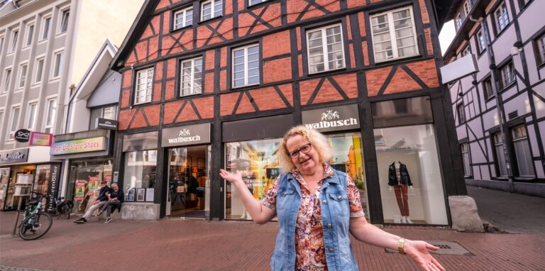 Eine blonde Frau steht vor einem Fachwerkhaus in Recklinghausen, in dessen Erdgeschoss ein Modeladen untergebracht ist.