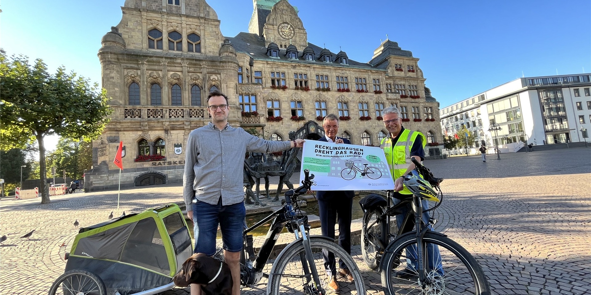 Drei Männer mit Fahrrädern halten zusammen ein Plakat hoch, auf dem „Recklinghausen dreht das Rad“ steht. Im Hintergrund ist das Recklinghäuser Rathaus zu sehen.