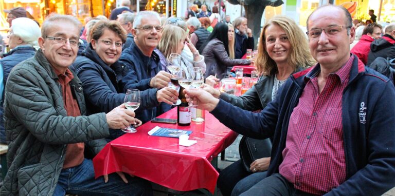 Männer und Frauen sitzen an einem Tisch und stoßen mit Weingläsern an.