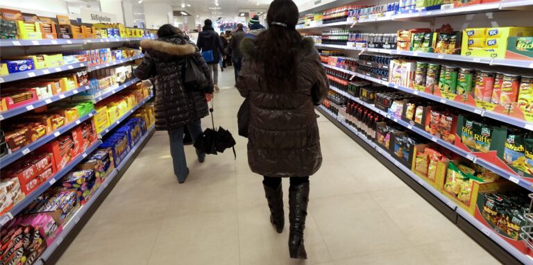 Kunden gehen durch einen Supermarkt-Gang. Rechts und links stehen Lebensmittelregale.